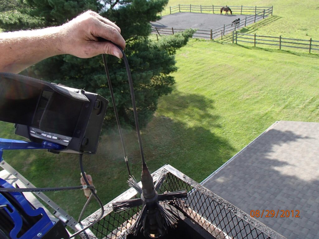 An aerial view from a rooftop showing a technician's hand lowering a chimney sweeping brush and camera probe into a flue with a diagnostic screen nearby, investigating chimney water intrusion in Louisville, KY.