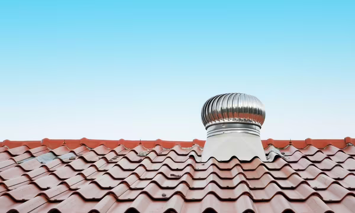 A silver metallic whirlybird turbine roof vent mounted on a red tile roof against a clear blue sky, requiring professional roof vent cleaning inspection services in Houston, TX.
