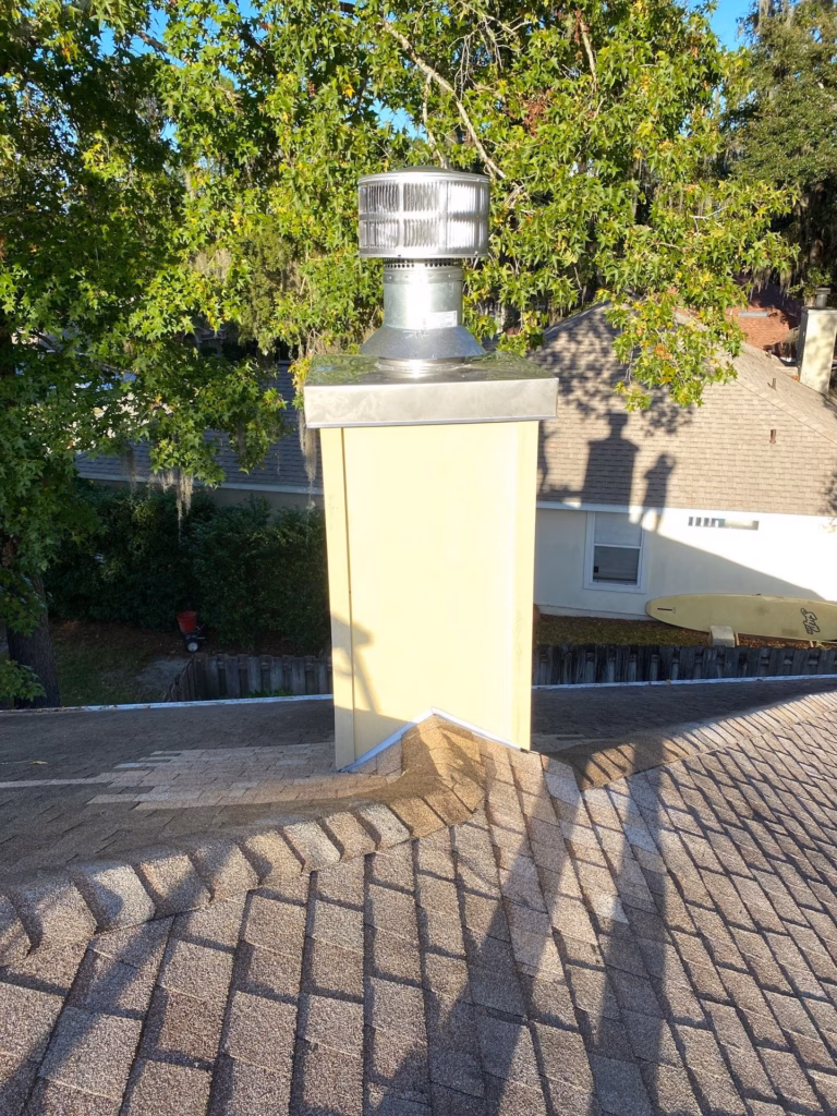 A pale yellow chimney chase featuring a newly installed metal chase cover and chimney cap spark arrestor on a shingled roof in Gainesville, FL.