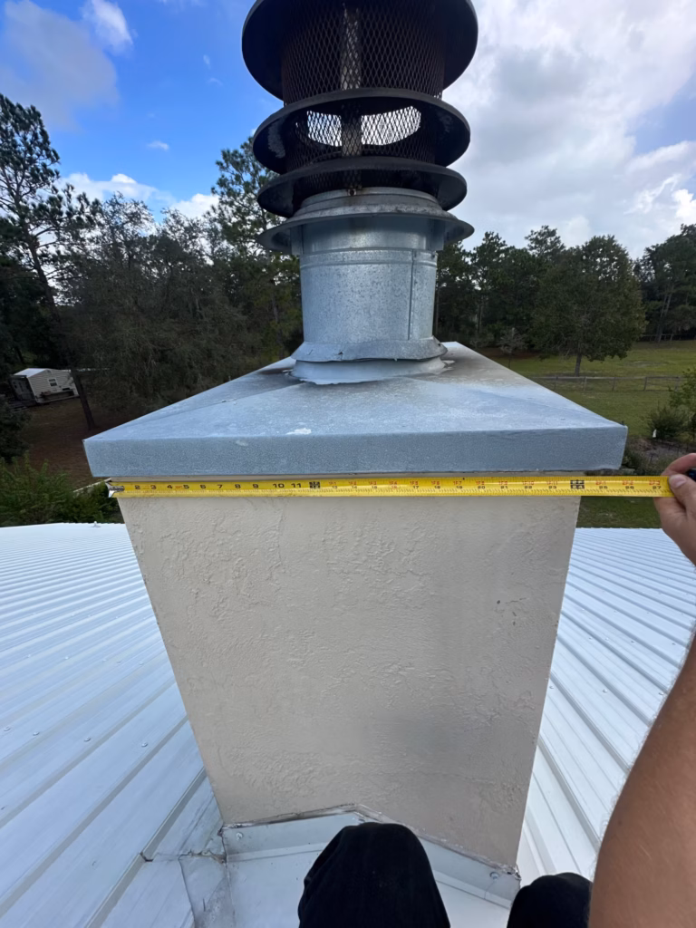 A technician measuring the top metal chase cover of a stucco chimney with a yellow tape measure on a metal roof in Gainesville, FL.
