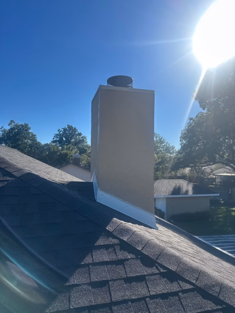 A newly repaired wood framed stucco chimney with freshly installed metal flashing and a round cap on a dark shingle roof under bright sunlight in Gainesville, FL.