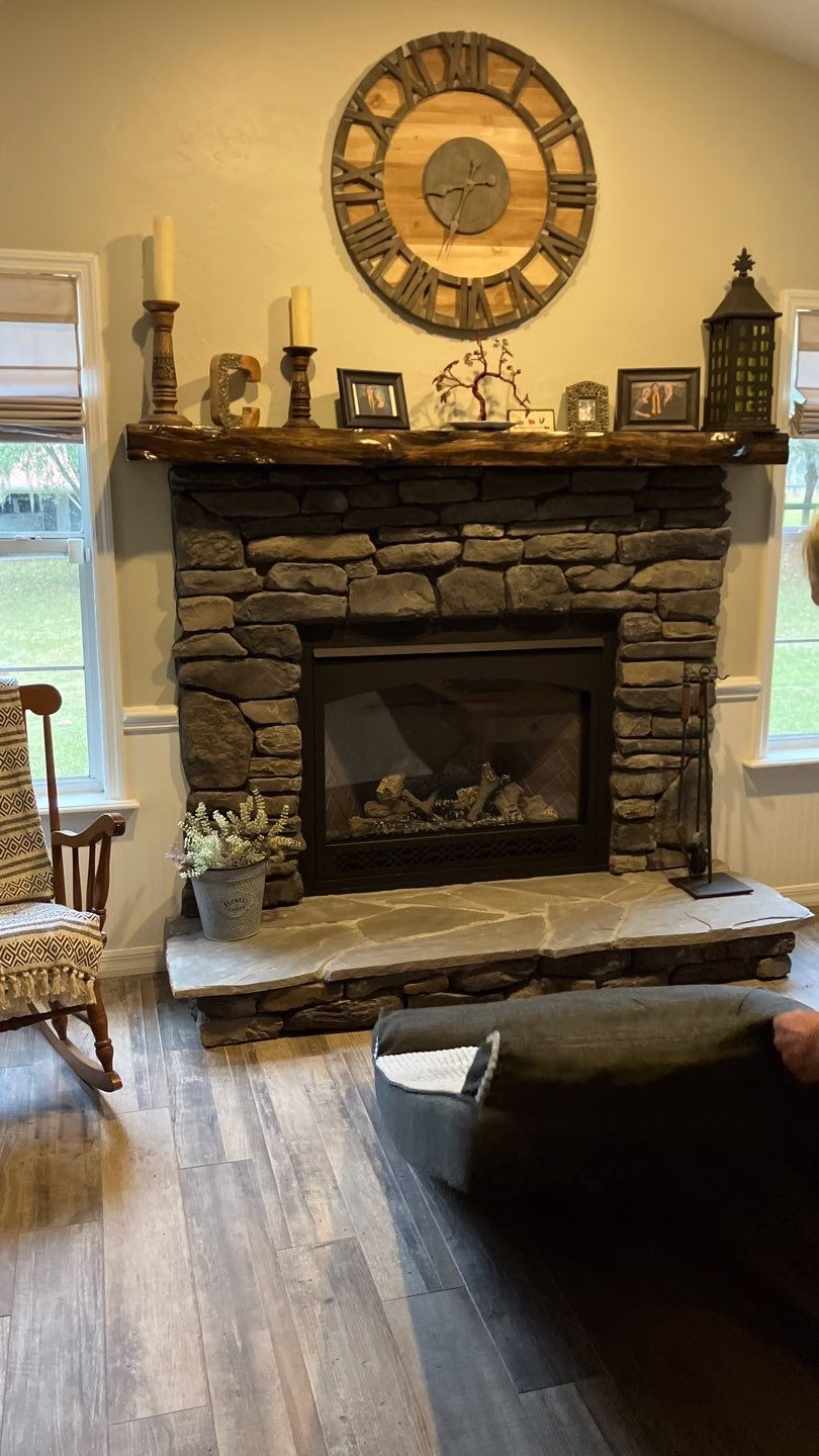 A cozy living room featuring a stacked stone fireplace with a live-edge wooden mantel, stone hearth, and decorative wall clock in Gainesville, FL.